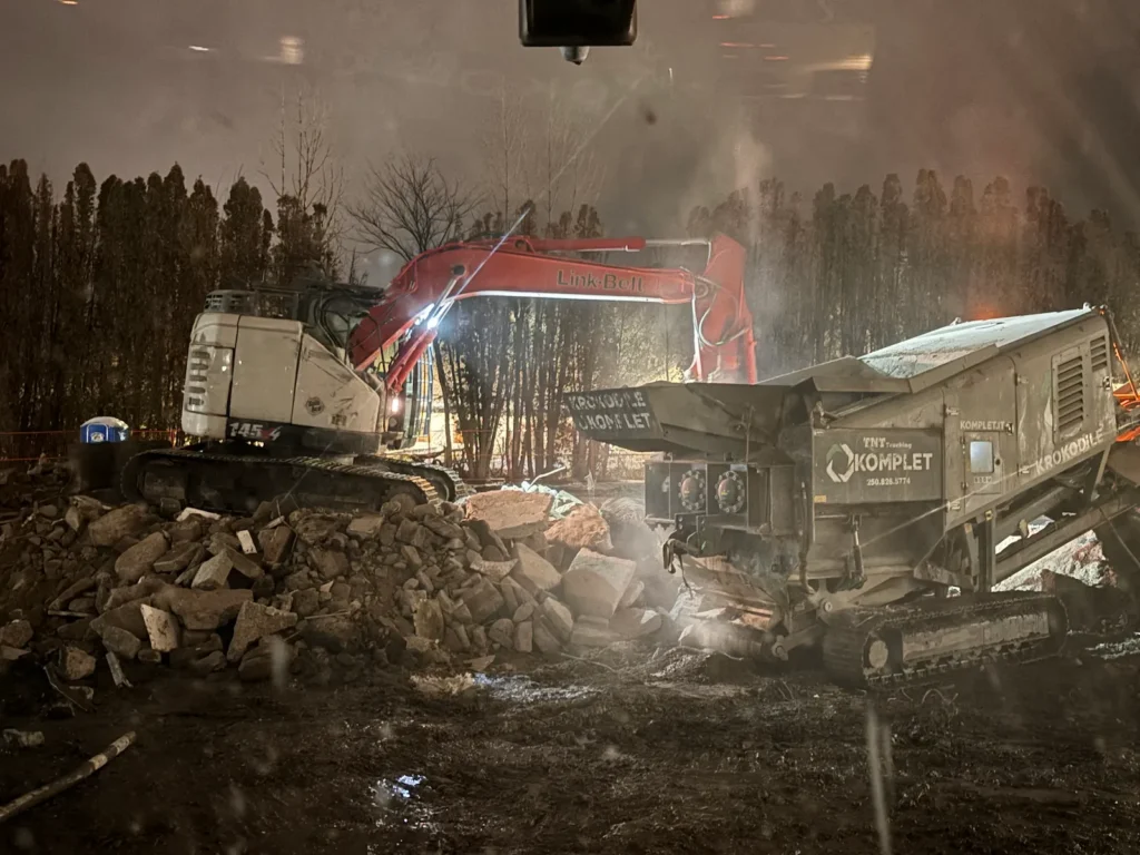 Excavator feeding a mobile crusher at night, processing concrete and rock on a muddy Kelowna jobsite.