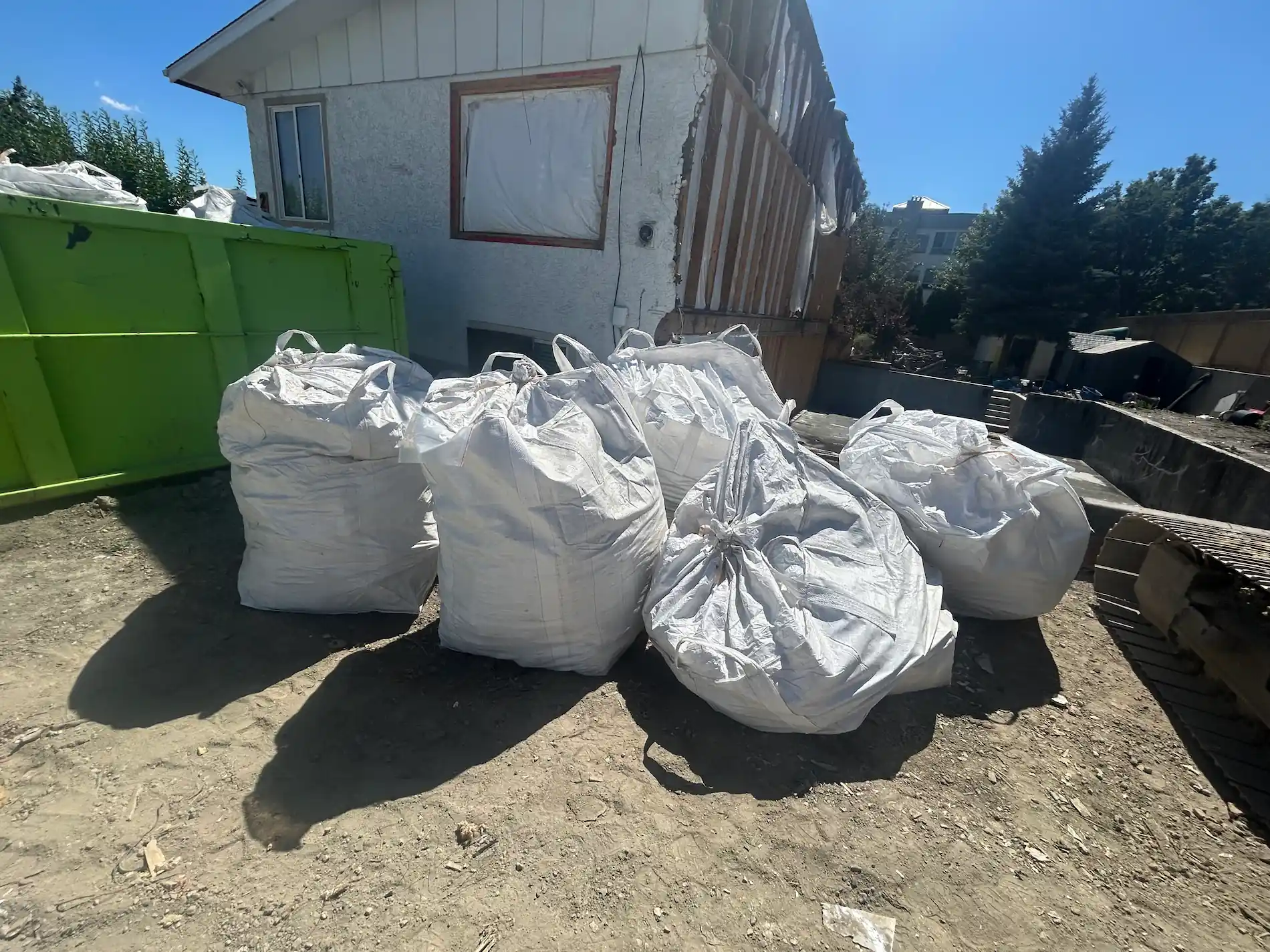 Bulk bags of construction debris beside a green dumpster at a residential renovation in Kelowna, ready for TNT junk removal pickup.